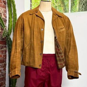 May include: A tan suede jacket with a pointed collar, long sleeves, and a button-front closure, worn over a plain white t-shirt and maroon athletic shorts. The jacket features a plaid lining in shades of green, red, and brown, with adjustable cuffs.