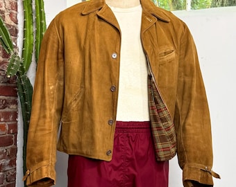 May include: A tan suede jacket with a pointed collar, long sleeves, and a button-front closure, worn over a plain white t-shirt and maroon athletic shorts. The jacket features a plaid lining in shades of green, red, and brown, with adjustable cuffs.
