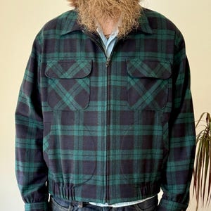 Könnte beinhalten: Eine grün-marineblau karierte Jacke mit Reißverschluss. Die Jacke hat zwei Vordertaschen und einen gerippten Saum. Die Ärmel haben geknöpfte Manschetten. Die Jacke wird über einem hellblauen Hemd und einer Jeans getragen.