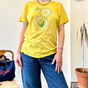 May include: Yellow t-shirt with a daisy and butterfly graphic, and the text "COOKIE CHAMP" in a circular design. The shirt is paired with blue jeans. A colorful beaded necklace and bracelet are also visible.