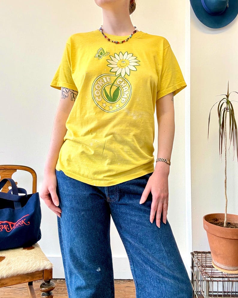 May include: Yellow t-shirt with a daisy and butterfly graphic, and the text "COOKIE CHAMP" in a circular design. The shirt is paired with blue jeans. A colorful beaded necklace and bracelet are also visible.
