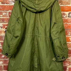 May include: An olive green parka coat featuring a large hood, long sleeves with buttoned cuffs, and a drawstring waist. The coat has a straight hemline with a central vent and visible stitching details throughout the fabric construction.