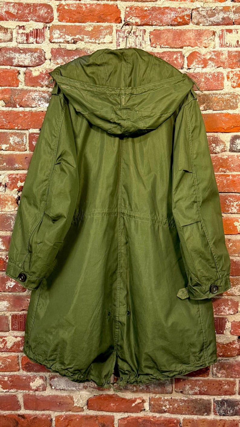 May include: An olive green parka coat featuring a large hood, long sleeves with buttoned cuffs, and a drawstring waist. The coat has a straight hemline with a central vent and visible stitching details throughout the fabric construction.
