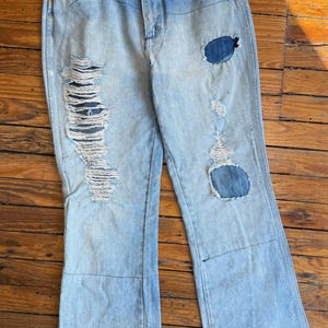 Op de afbeelding: Lichtblauwe spijkerbroek met distressed details. De jeans heeft gescheurde delen aan de voorkant van de benen en patches. De jeans heeft een klassiek ontwerp met vijf zakken en een uitlopende beenopening.