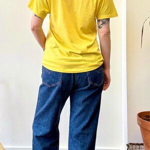 May include: A person wearing a yellow t-shirt and dark blue jeans. The jeans have red stitching on the back pockets. The person is also wearing turquoise and gray sneakers. The background is a plain white wall.