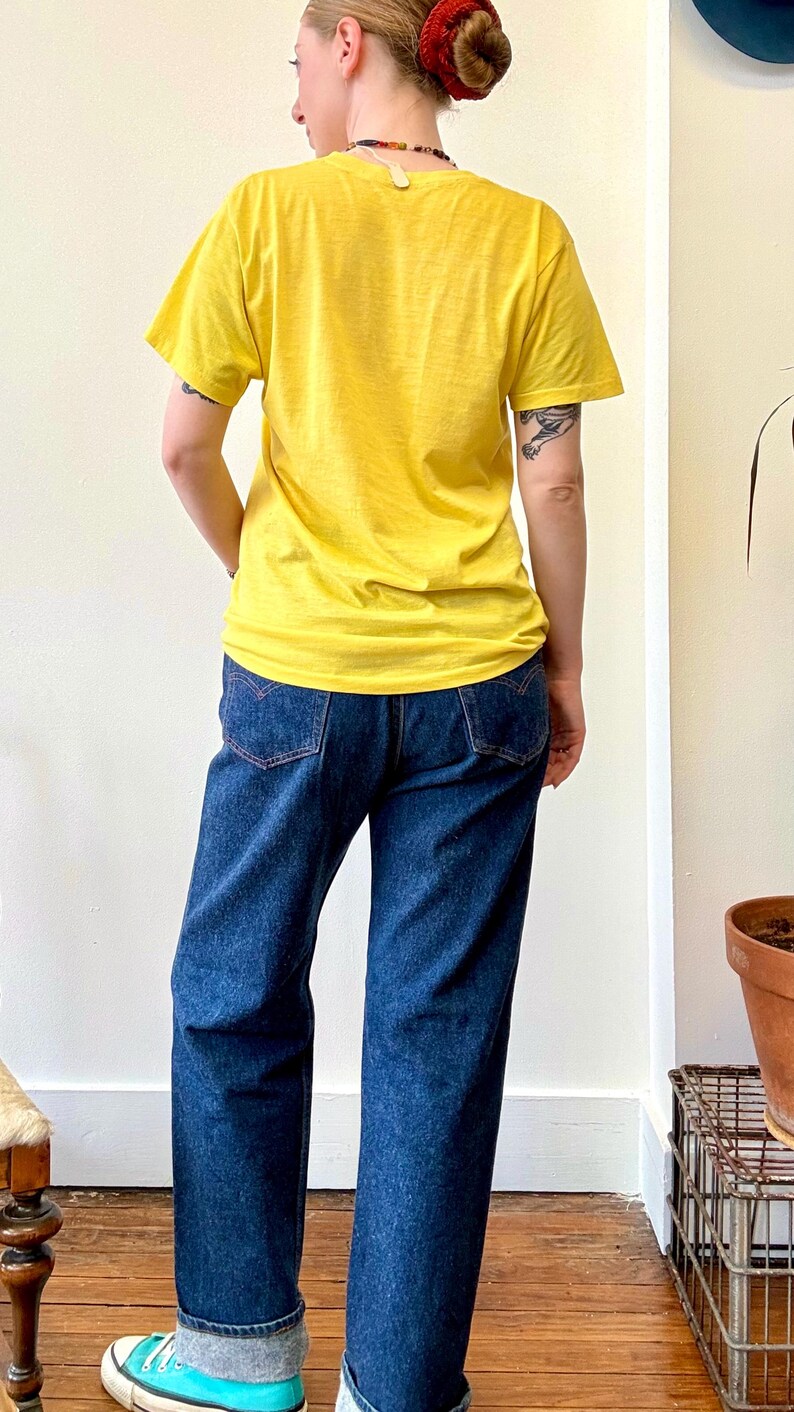 May include: A person wearing a yellow t-shirt and dark blue jeans. The jeans have red stitching on the back pockets. The person is also wearing turquoise and gray sneakers. The background is a plain white wall.
