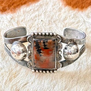 May include: A silver cuff bracelet with a rectangular, reddish-brown stone with black and white patterns. The stone is framed by a beaded border. Two silver, rounded accents flank the central stone.
