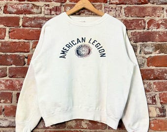 Puede incluir: Sudadera de cuello redondo color crema con la inscripción "AMERICAN LEGION" en azul marino arqueada sobre el pecho. El texto tiene un emblema circular en el centro. La sudadera cuelga de una percha de madera contra una pared de ladrillos.
