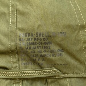 May include: An olive green parka shell featuring a printed label with the text PARKA-SHELL, RA-JEF MFG CO., 23462-01-8650, JANUARY 1952, NO. MIL-P-11013 QMC, DATE MARCH 12, 1951, N.M.P.A., INSPECTOR. The garment includes visible stitching and a button.