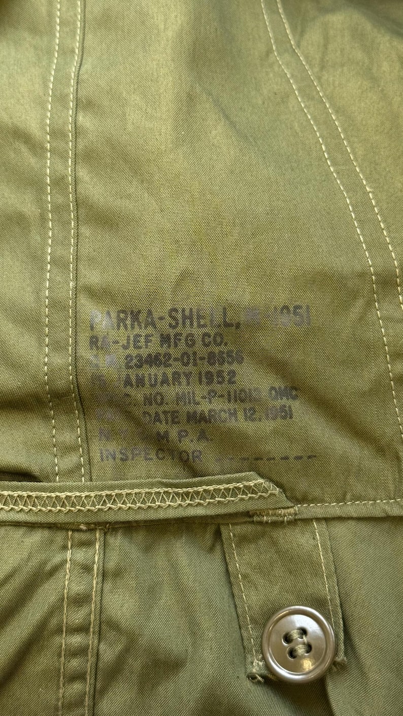 May include: An olive green parka shell featuring a printed label with the text PARKA-SHELL, RA-JEF MFG CO., 23462-01-8650, JANUARY 1952, NO. MIL-P-11013 QMC, DATE MARCH 12, 1951, N.M.P.A., INSPECTOR. The garment includes visible stitching and a button.