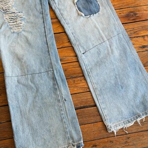 Op de afbeelding: Lichtblauwe denim jeans met distressed details, waaronder gescheurde delen en patches. De jeans hebben een wijd been ontwerp en gerafelde zomen, waardoor een vintage, versleten look ontstaat. De jeans worden getoond op een houten oppervlak.