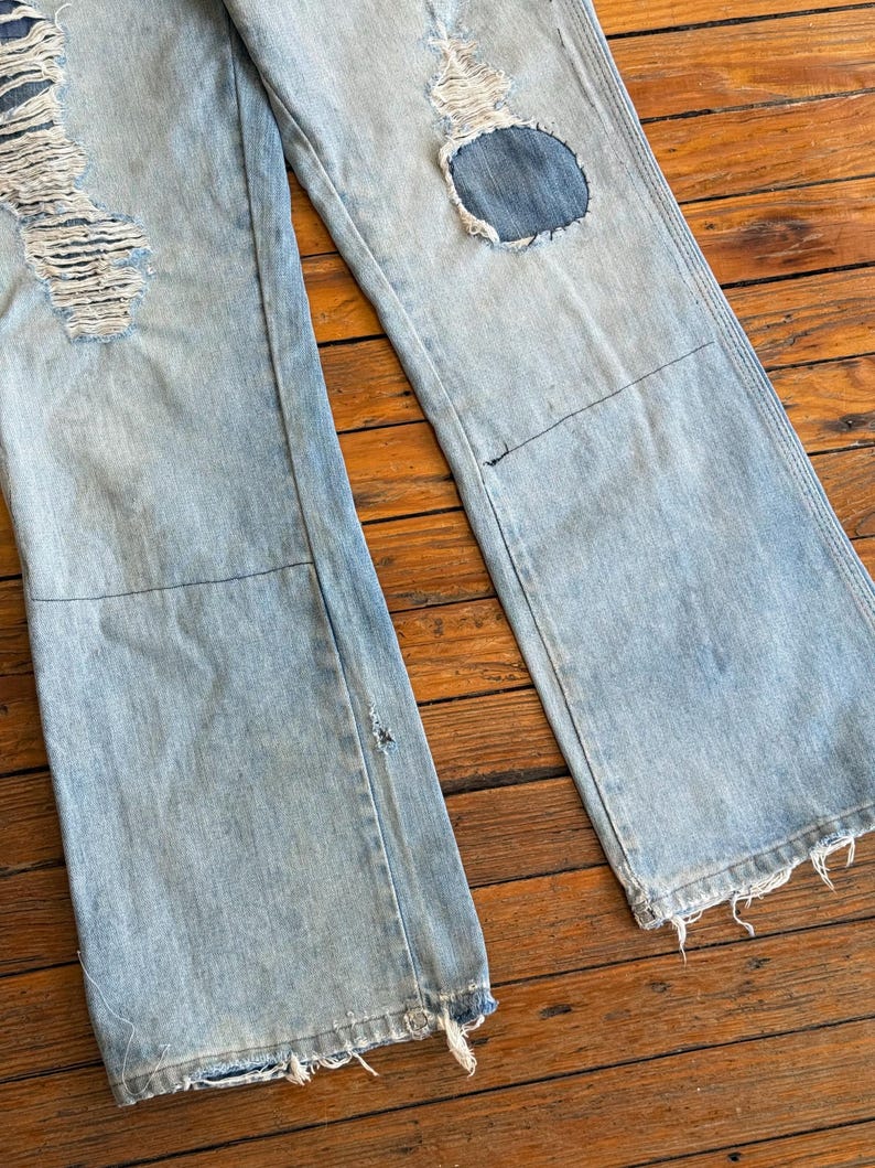 Op de afbeelding: Lichtblauwe denim jeans met distressed details, waaronder gescheurde delen en patches. De jeans hebben een wijd been ontwerp en gerafelde zomen, waardoor een vintage, versleten look ontstaat. De jeans worden getoond op een houten oppervlak.