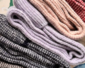 Op de afbeelding: Een stapel van vier paar gebreide sokken in verschillende kleuren en patronen. De sokken zijn gevouwen en gestapeld, met de bovenste sok in een licht roze kleur, gevolgd door een licht paarse sok, een zwart-wit gepatroneerde sok en een donkergrijze sok.