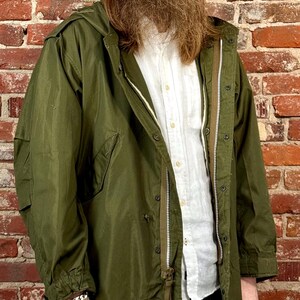 May include: An olive green hooded parka jacket featuring a full-length front zipper, a button-down placket, long sleeves with adjustable cuffs, and two large side pockets. The jacket is worn over a white button-up shirt and dark blue denim jeans.