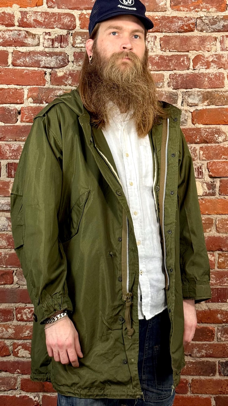 May include: An olive green hooded parka jacket featuring a full-length front zipper, a button-down placket, long sleeves with adjustable cuffs, and two large side pockets. The jacket is worn over a white button-up shirt and dark blue denim jeans.