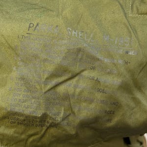 May include: An olive green parka shell M-1951 with a hood and a single visible button closure. The interior fabric features printed text including 'PARKA SHELL M-1951' and instructions regarding garment care, zipper lubrication, and heat exposure.