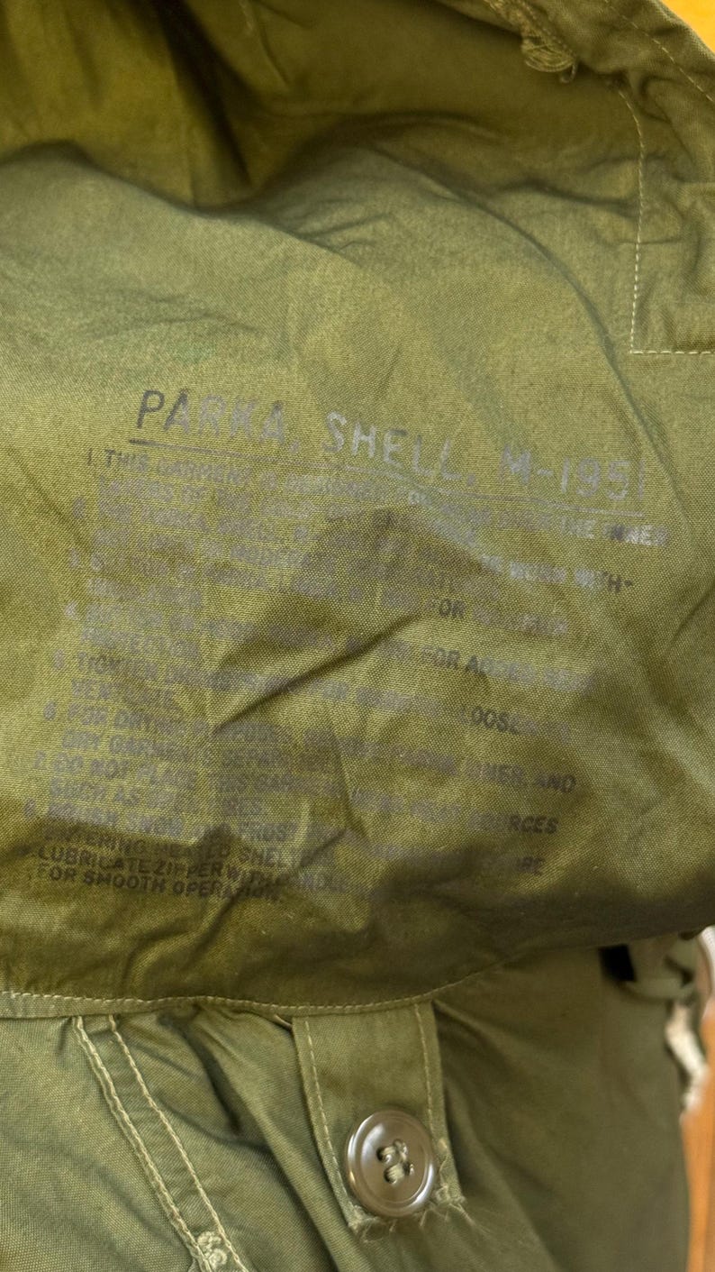 May include: An olive green parka shell M-1951 with a hood and a single visible button closure. The interior fabric features printed text including 'PARKA SHELL M-1951' and instructions regarding garment care, zipper lubrication, and heat exposure.