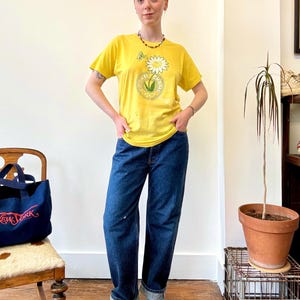 May include: A person wearing a yellow t-shirt with a daisy graphic and the text "Cookie Champs". The person is also wearing blue jeans and teal sneakers. A navy blue tote bag with "New York" text is visible.