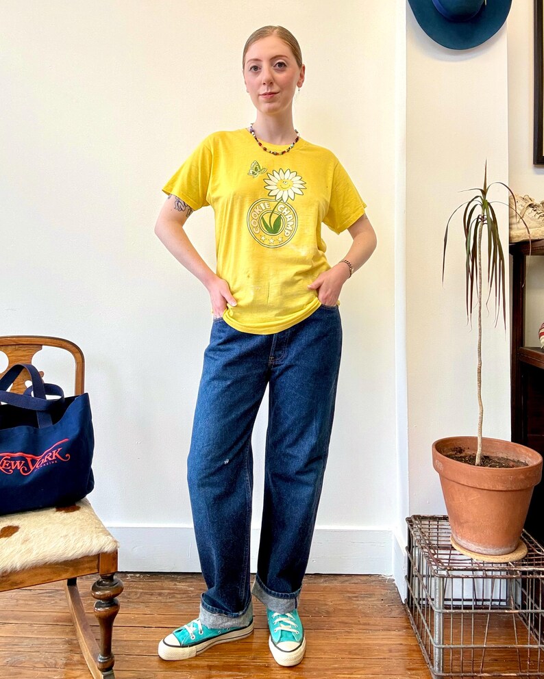 May include: A person wearing a yellow t-shirt with a daisy graphic and the text "Cookie Champs". The person is also wearing blue jeans and teal sneakers. A navy blue tote bag with "New York" text is visible.