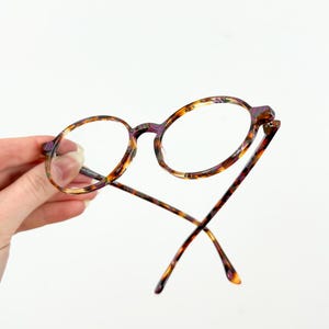 May include: Round tortoiseshell eyeglasses with purple accents. The glasses are held in a hand and the arms are crossed.