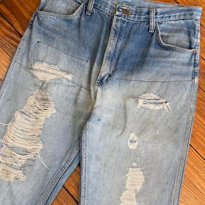 May include: Light blue denim jeans with distressed details, including ripped sections and visible stitching. The jeans have a classic five-pocket design and a button and zipper closure. The denim has a faded appearance.