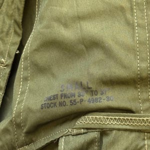 May include: An olive green fabric garment featuring visible stitching and a button closure. Printed text on the fabric reads: SMALL, CHEST FROM 33 TO 37, STOCK NO. 55-P-4982-30. The material has a textured weave and includes a reinforced seam detail.