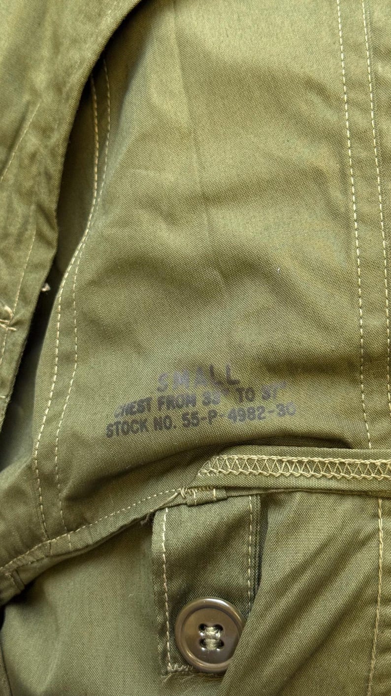 May include: An olive green fabric garment featuring visible stitching and a button closure. Printed text on the fabric reads: SMALL, CHEST FROM 33 TO 37, STOCK NO. 55-P-4982-30. The material has a textured weave and includes a reinforced seam detail.