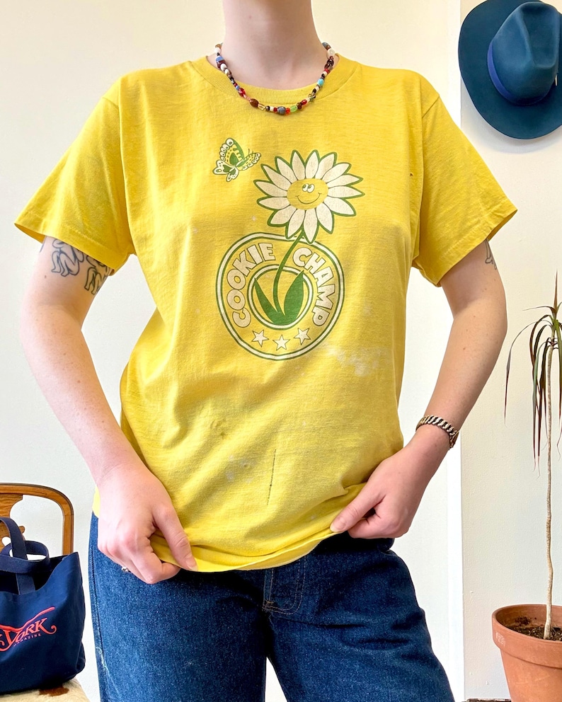 May include: Yellow t-shirt with a daisy graphic and the text "COOKIE CHAMP". The shirt also features a butterfly graphic. The shirt is paired with blue jeans.