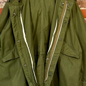 May include: An olive green military-style parka featuring a full-length front zipper, a button-down storm flap, a hood, and two large front pockets. The jacket is constructed with visible stitching and includes internal drawstrings at the waist.