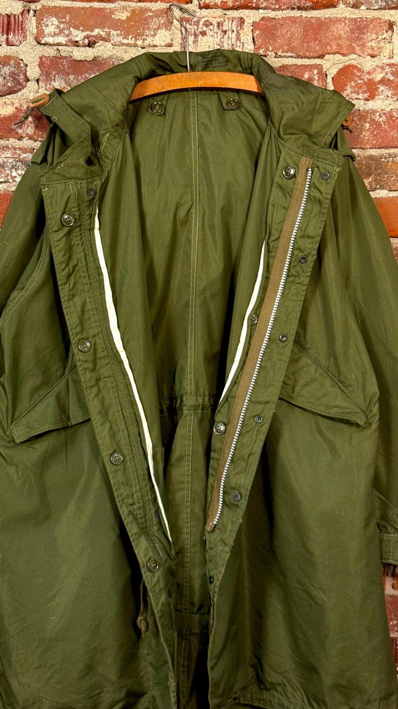 May include: An olive green military-style parka featuring a full-length front zipper, a button-down storm flap, a hood, and two large front pockets. The jacket is constructed with visible stitching and includes internal drawstrings at the waist.
