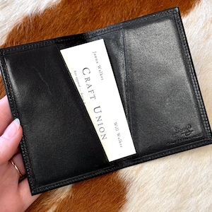 May include: Black leather wallet with a card that reads "CRAFT UNION" and the names "Janna Walker" and "Will Walker". The wallet is open, revealing a pocket. The background is a brown and white cowhide pattern.
