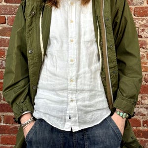 May include: An olive green field jacket with a front zipper and button closure, worn over a white button-down shirt and dark blue denim shorts. The jacket features long sleeves and a hood, while the shorts have large front pockets.