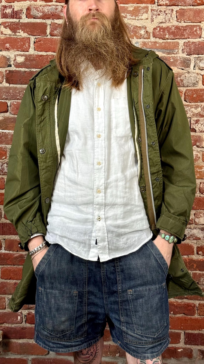 May include: An olive green field jacket with a front zipper and button closure, worn over a white button-down shirt and dark blue denim shorts. The jacket features long sleeves and a hood, while the shorts have large front pockets.