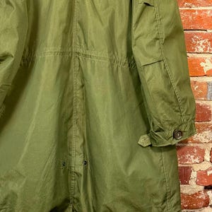 May include: An olive green hooded parka featuring a long silhouette, a drawstring waist, and long sleeves with buttoned cuffs. The garment includes a rear fishtail hem design with visible snap closures and reinforced stitching throughout the fabric.
