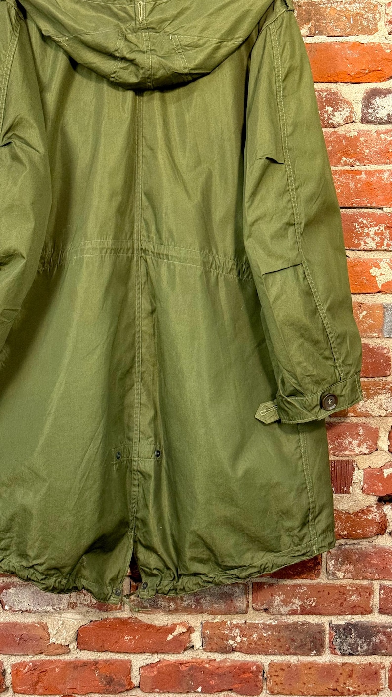 May include: An olive green hooded parka featuring a long silhouette, a drawstring waist, and long sleeves with buttoned cuffs. The garment includes a rear fishtail hem design with visible snap closures and reinforced stitching throughout the fabric.