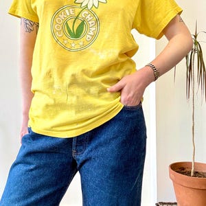 May include: Yellow t-shirt with a daisy and butterfly graphic, and the text "COOKIE CHAMP" in a circular design. The shirt is paired with blue jeans. A colorful beaded necklace and bracelet are also visible.