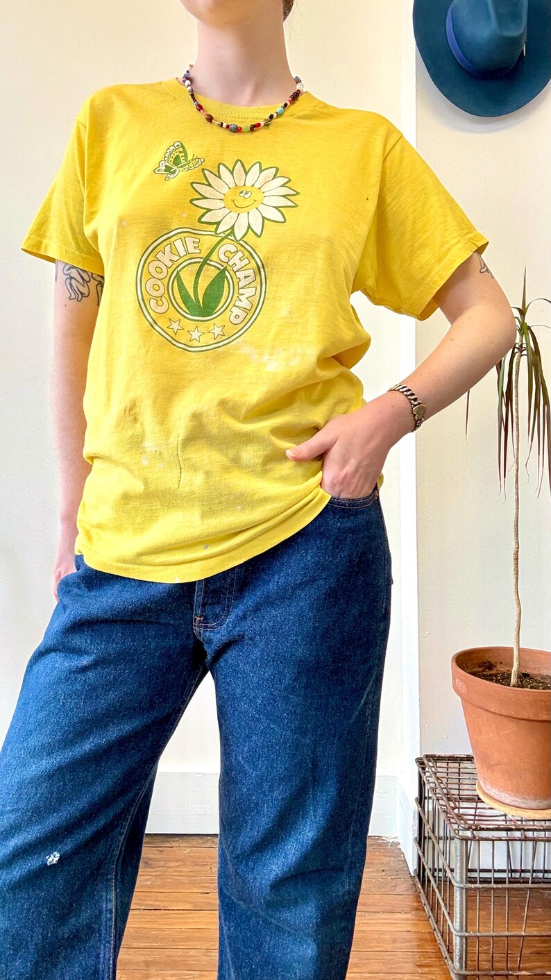 May include: Yellow t-shirt with a daisy and butterfly graphic, and the text "COOKIE CHAMP" in a circular design. The shirt is paired with blue jeans. A colorful beaded necklace and bracelet are also visible.