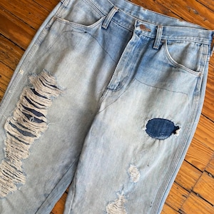 Op de afbeelding: Lichtblauwe denim jeans met distressed details, waaronder grote scheuren en patches. De jeans hebben een klassiek ontwerp met vijf zakken en een knoop- en ritssluiting. De denim heeft een vervaagde wassing.