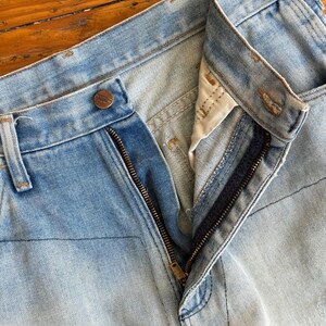 Op de afbeelding: Close-up van een lichtblauwe spijkerbroek met een gedeeltelijk geopende ritssluiting. De jeans heeft een knoopsluiting en zichtbare stikseldetails. De denim heeft een vervaagde uitstraling.