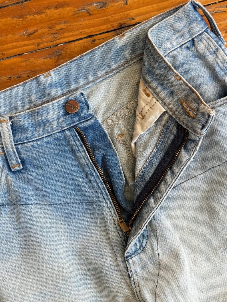 Op de afbeelding: Close-up van een lichtblauwe spijkerbroek met een gedeeltelijk geopende ritssluiting. De jeans heeft een knoopsluiting en zichtbare stikseldetails. De denim heeft een vervaagde uitstraling.