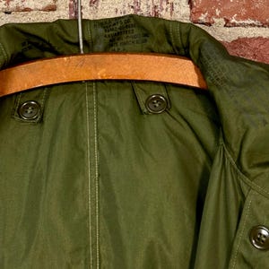 May include: An olive green military-style parka jacket featuring a high collar, two dark buttons on the front placket, and a wooden hanger. The interior neck area displays printed text including the date January 1952 and military specifications.