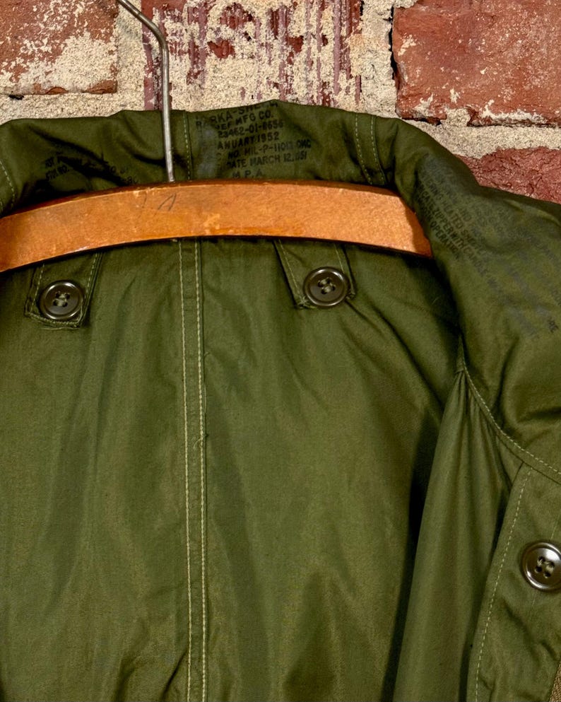 May include: An olive green military-style parka jacket featuring a high collar, two dark buttons on the front placket, and a wooden hanger. The interior neck area displays printed text including the date January 1952 and military specifications.