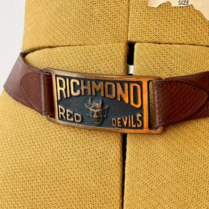 May include: Brown leather belt with a rectangular buckle. The buckle features the words "RICHMOND RED DEVILS" and a devil face. The belt is displayed on a yellow mannequin.