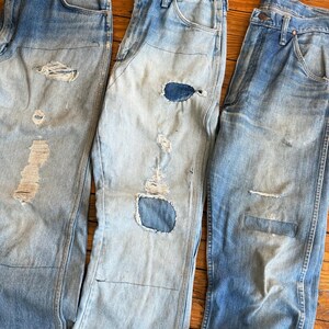 Op de afbeelding: Drie spijkerbroeken van lichtblauwe denim met distressed details. Elke broek heeft scheuren en patches, wat een vintage of versleten uitstraling laat zien. De jeans hebben een klassieke vijf-zakken-stijl en een vervaagde wassing.