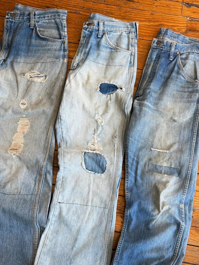 Op de afbeelding: Drie spijkerbroeken van lichtblauwe denim met distressed details. Elke broek heeft scheuren en patches, wat een vintage of versleten uitstraling laat zien. De jeans hebben een klassieke vijf-zakken-stijl en een vervaagde wassing.