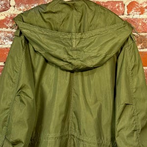 May include: An olive green hooded parka jacket featuring a large, structured hood with visible stitching, long sleeves, and a central vertical seam running down the back. The garment is constructed from a solid-colored fabric with a smooth texture.