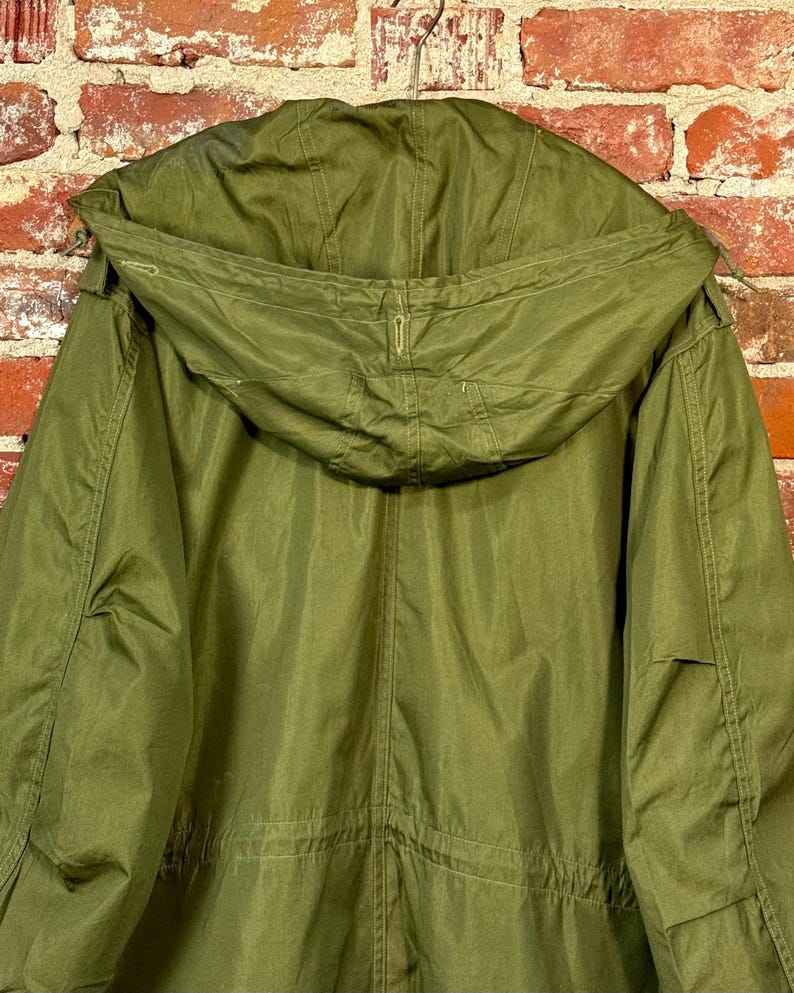 May include: An olive green hooded parka jacket featuring a large, structured hood with visible stitching, long sleeves, and a central vertical seam running down the back. The garment is constructed from a solid-colored fabric with a smooth texture.