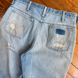 Op de afbeelding: Lichtblauwe denim jeans met distressed details, waaronder scheuren en versleten patches. De achterzakken hebben het Wrangler-logo en een kenmerkend stikselontwerp. De jeans zijn vintage en vertonen gebruikssporen.