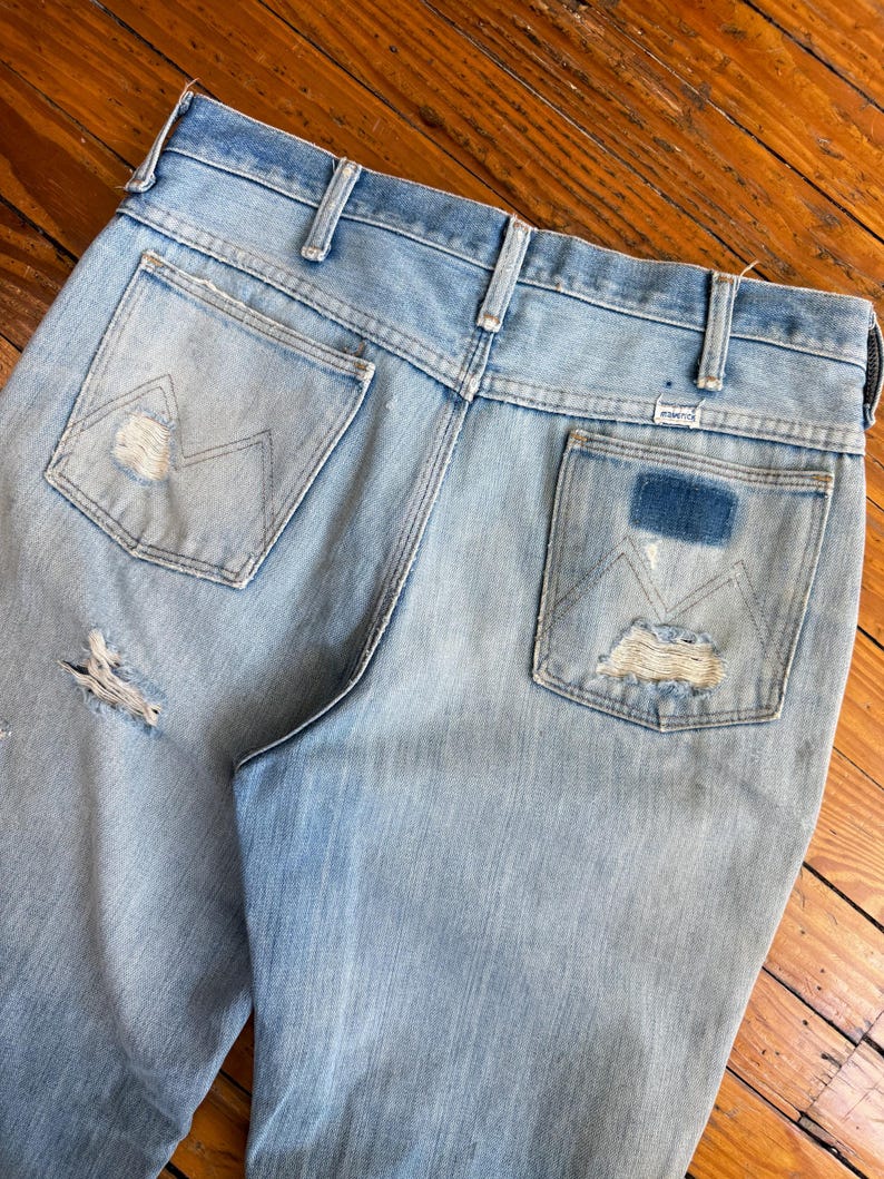 Op de afbeelding: Lichtblauwe denim jeans met distressed details, waaronder scheuren en versleten patches. De achterzakken hebben het Wrangler-logo en een kenmerkend stikselontwerp. De jeans zijn vintage en vertonen gebruikssporen.