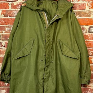 May include: An olive green military-style parka featuring a large hood, a front zipper closure concealed by a snap-button placket, and two large flap pockets on the front. The sleeves have adjustable cuffs with buttons and the hood has a drawstring.